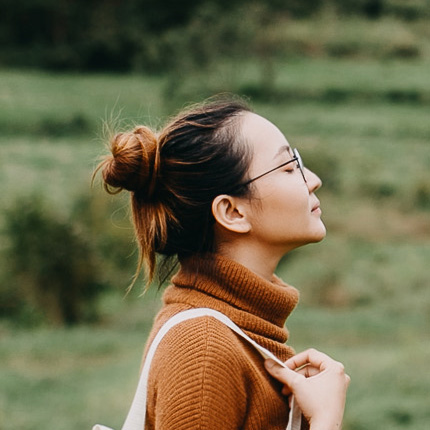 A side profile of a woman in a russet-coloured turtleneck and white bag. She looks up with her eyes closed.